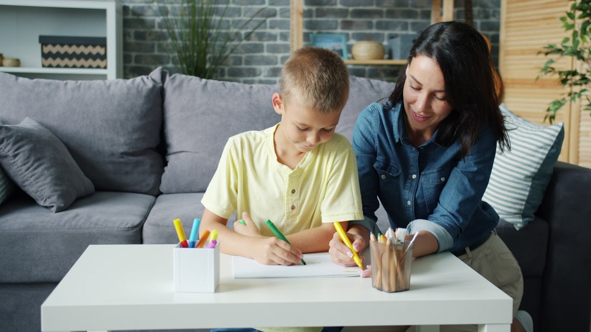 Small boy is drawing with markers together with caring mother in cozy room at home enjoying activity making picture. Creative kids and loving parents concept.